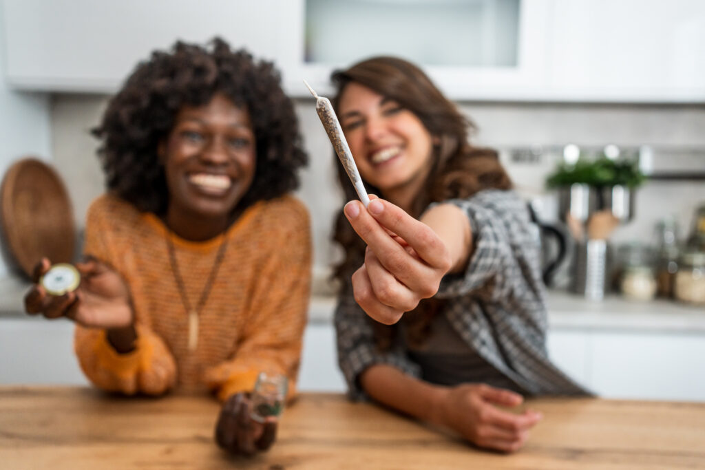 Two young women smiling while holding a cannabis joint and medical marijuana in a cozy kitchen, enjoying moments of friendship and relaxation